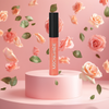 A single lip gloss tube stands upright on a round pink pedestal against a pink backdrop with floating peach roses and green leaves. The tube has a black cap and a peachy pink translucent gloss inside, with the brand name MOECHA//ADE printed vertically on the front.