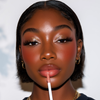Close-up of a model with dark hair and warm-toned makeup applying a glossy nude lip product with a wand applicator.