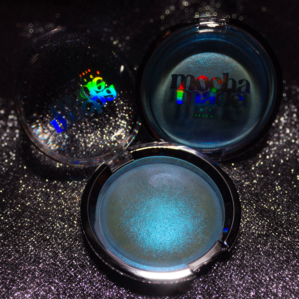 Three round makeup compacts on a sparkly black background. One compact is open, showing a shimmering light blue pressed powder.