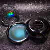 An open round makeup compact showing a shimmering light blue-green pressed powder, with the black lid placed beside it on a sparkly dark background. The lid is printed with the brand name 'Mermaid Money' and 'YG'.