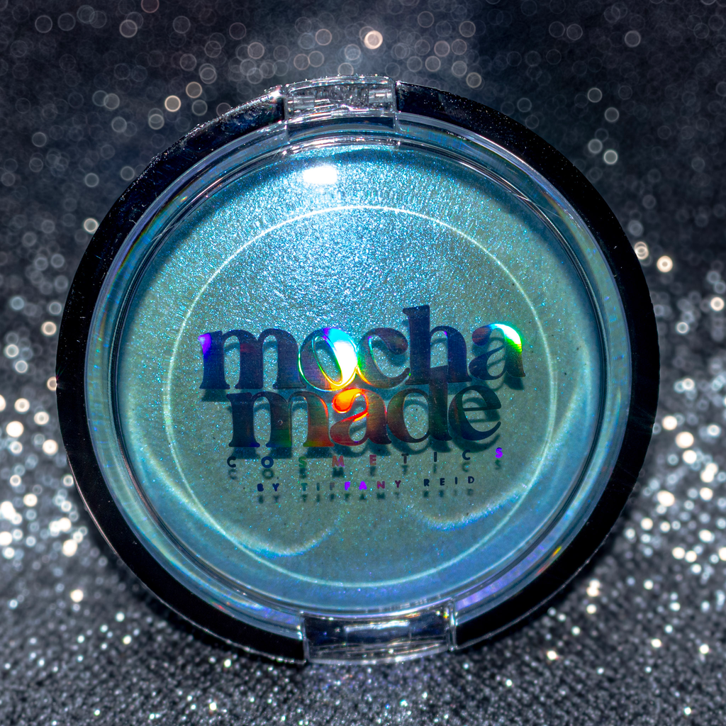 A round makeup compact with a black rim and a clear lid showing shimmering blue-green eyeshadow. The lid has holographic text reading 'mocha made cosmetics by Jennifer Auld' on a sparkly dark background.