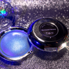 An open round makeup compact showing a shimmering blue-purple pressed powder labeled 'Mermaid Money,' with the black lid placed beside it on a sparkly silver surface.