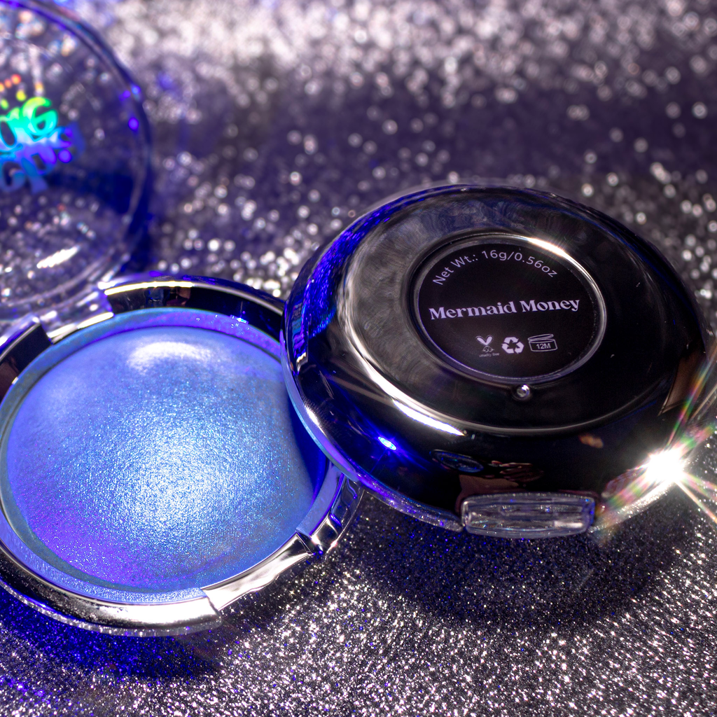 An open round makeup compact showing a shimmering blue-purple pressed powder labeled 'Mermaid Money,' with the black lid placed beside it on a sparkly silver surface.
