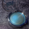 An open round makeup compact showing a shimmering light blue eyeshadow or pressed powder, with the translucent lid partly visible and the brand name appearing on the inside.