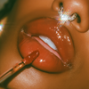 Close-up of glossy warm brown lips wearing a matching lip product, with a lip applicator touching the lower lip and a sparkling rhinestone nose ring visible against warm, high-contrast lighting.