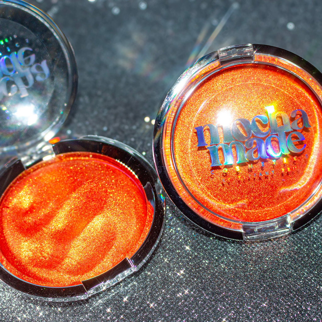 Shimmering molten tangerine golden chrome highlighter makeup compact with 'mocha made cosmetics' branding on a glittery background