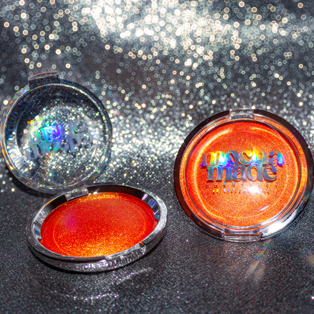 Shimmering molten tangerine golden chrome highlighter makeup compact with 'mocha made cosmetics' branding on a glittery background