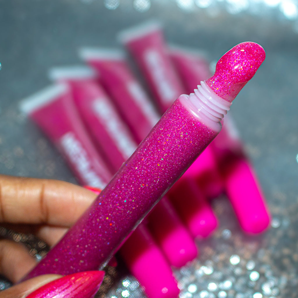 A close-up of a hand holding a tube of glittery pink lip gloss, with several matching pink lip gloss tubes blurred in the background on a sparkly surface.