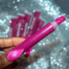 A close-up of a hand holding a tube of glittery pink lip gloss or liquid lipstick, with several matching tubes blurred in the background on a sparkly silver surface.