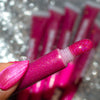 A close-up of a hand holding a tube of glittery pink lip gloss or gel, with the applicator tip coated in sparkling pink product. The background is a blurred silver glitter texture with more matching pink tubes visible.
