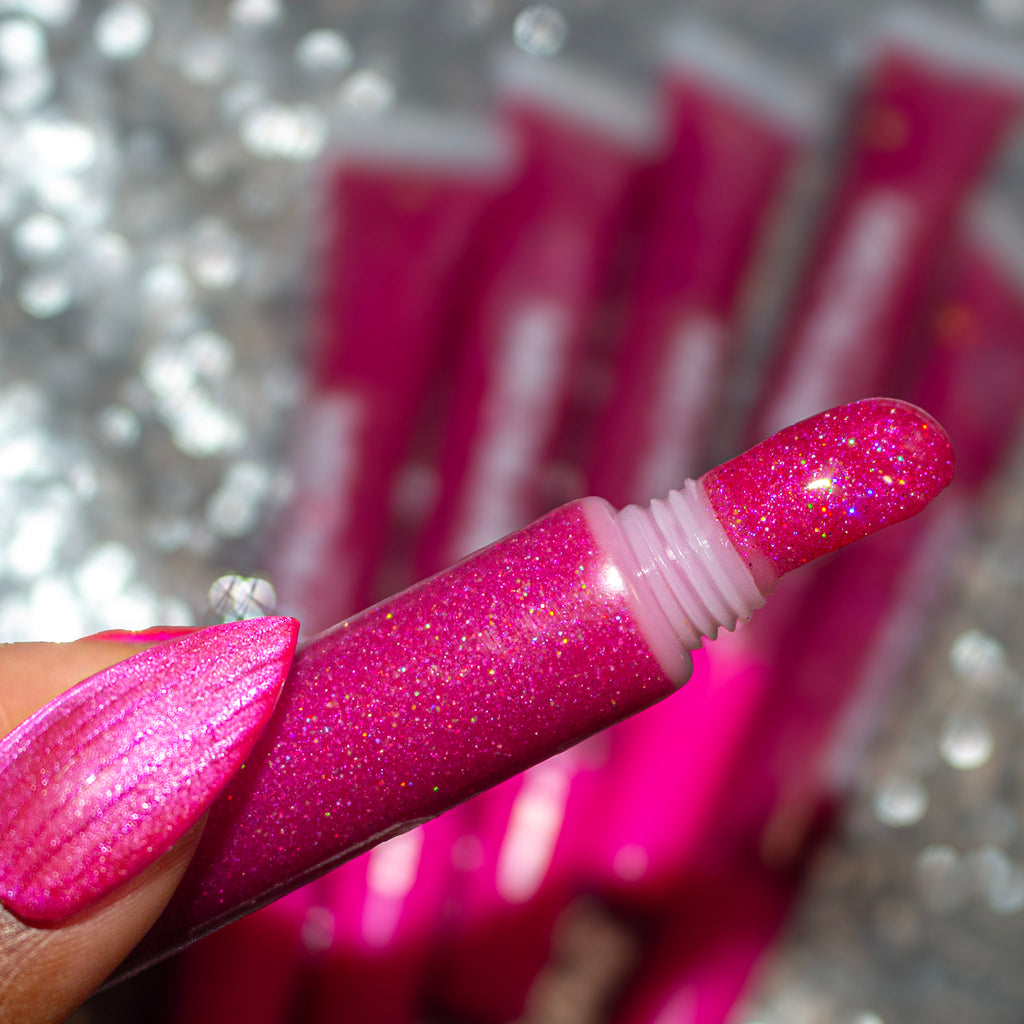 A close-up of a hand holding a tube of glittery pink lip gloss or gel, with the applicator tip coated in sparkling pink product. The background is a blurred silver glitter texture with more matching pink tubes visible.