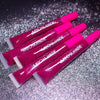 Four tubes of Mocha Made glitter lip gloss in a vibrant magenta-pink shade, arranged diagonally on a sparkly silver background.