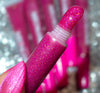 A close-up of a pink glitter lip gloss tube held in a hand with matching pink glitter nails, with more pink tubes blurred in the background.
