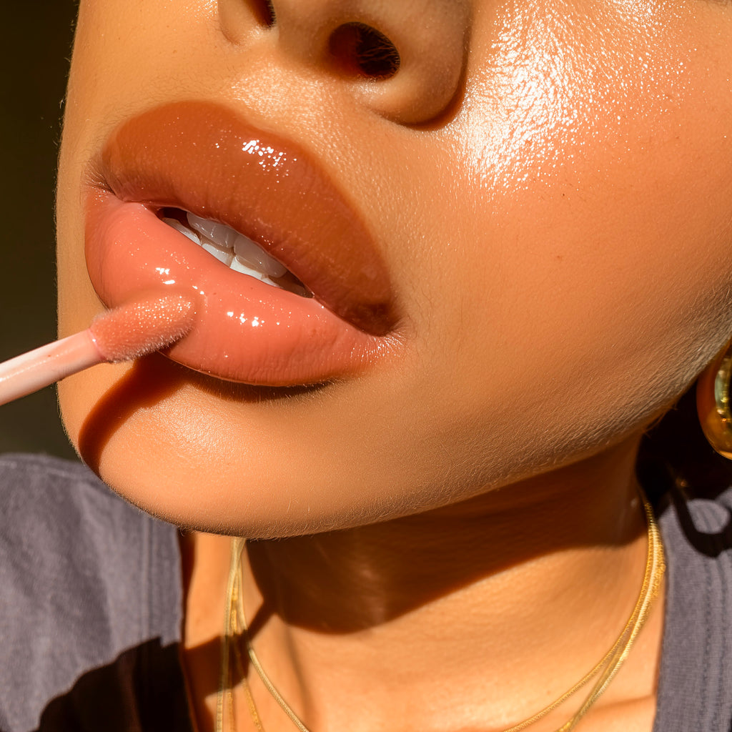 Close-up of a person applying a glossy peach-pink lip gloss with a doe-foot applicator, showing the product on the lips against warm sunlight.