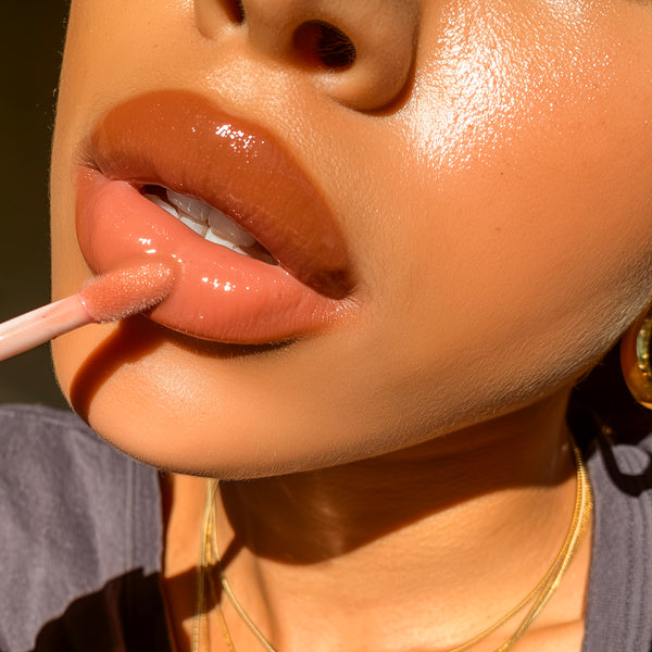 Close-up of a person applying a glossy peach-pink lip gloss with a doe-foot applicator, showing the product on the lips against warm sunlight.