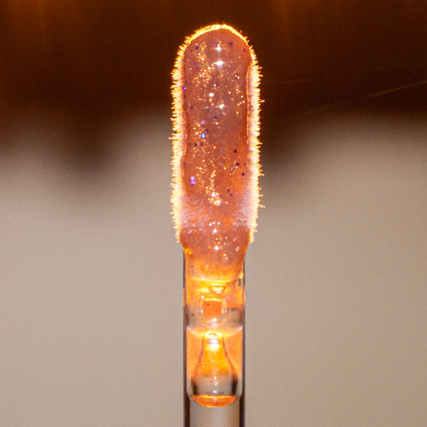 A close-up of a clear cosmetic dropper or pipette containing glowing amber-orange liquid with fine glitter and sparkly flecks, set against a softly blurred warm brown and beige background.