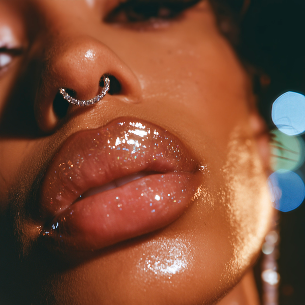 Extreme close-up of a person's face highlighting glossy, sparkly nude-pink lips and a curved rhinestone nose ring, with warm dramatic lighting and blurred blue bokeh in the background.
