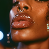 Close-up of glossy, sparkly nude-pink lips with a rhinestone nose ring and warm-toned skin under dramatic lighting.