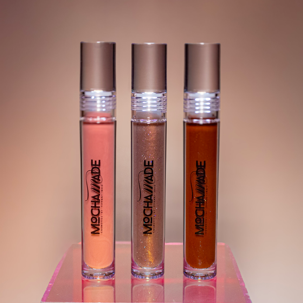 Three lip gloss tubes from Mocha Made Cosmetics on a reflective surface with a gradient background.