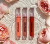 Three lip gloss tubes from Mocha Made Cosmetics on a reflective surface with a gradient background.